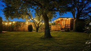 Una cabaña de madera con un gran árbol en primer plano, rodeada de un césped cuidado y una valla de bambú.