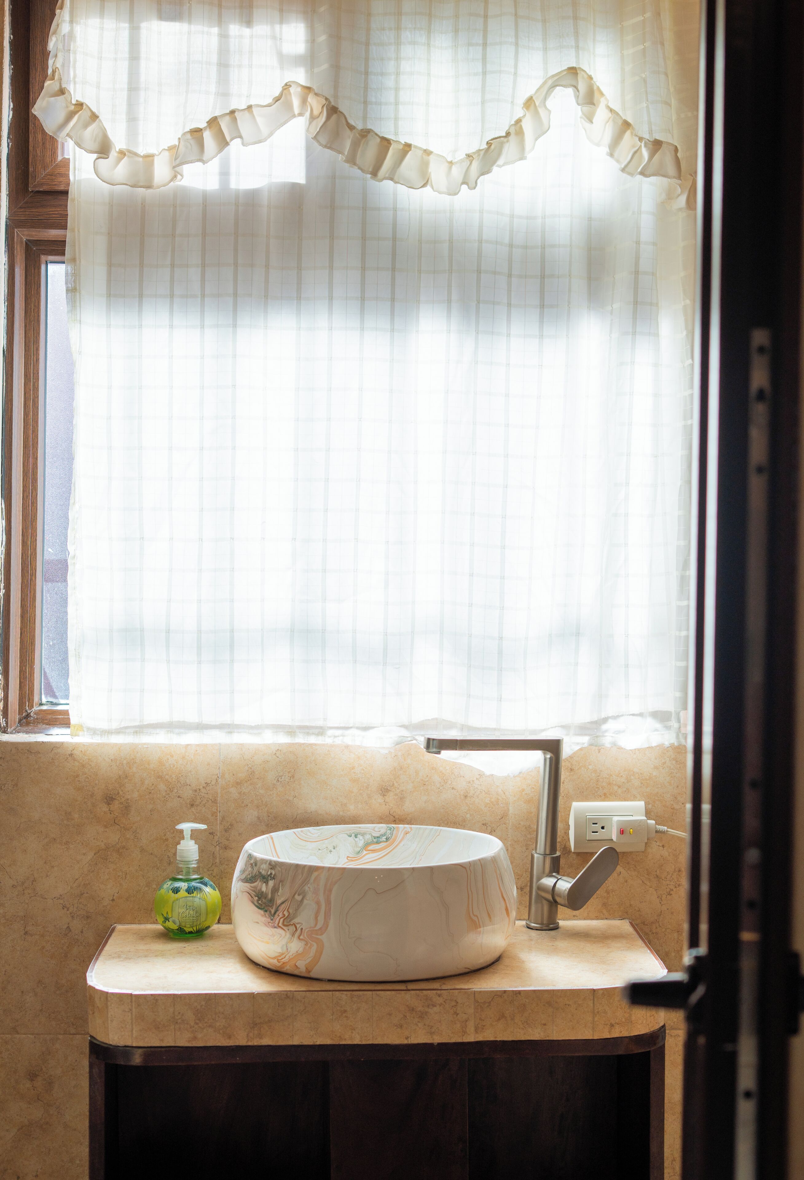 A bathroom with a marble sink, a soap dispenser, and a window with sheer curtains.
