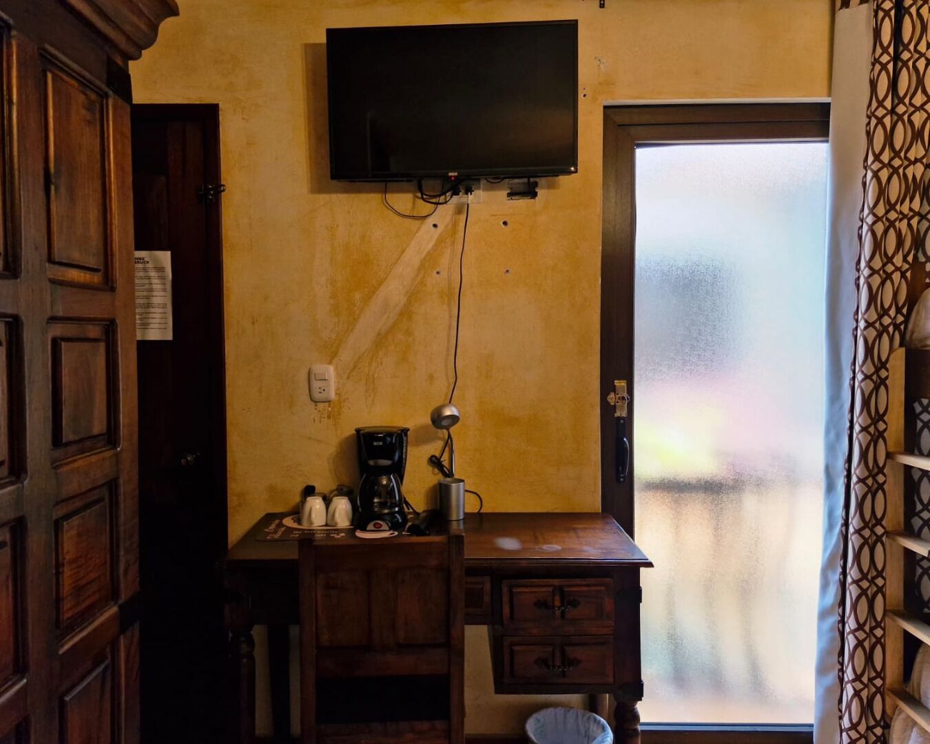 A small room with a wooden desk, a coffee maker, a television mounted on the wall, and a door leading to a frosted glass window.