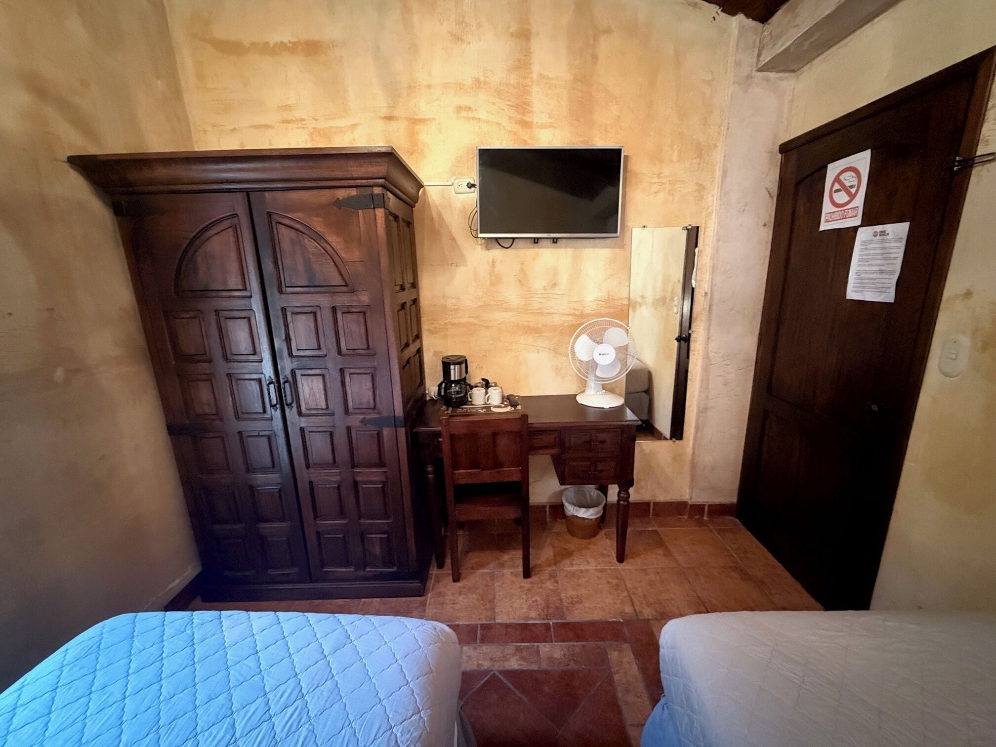 A small room with two beds, a wooden wardrobe, a desk with a fan, and a TV mounted on the wall.