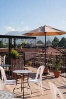 Un patio al aire libre con mesas y sillas, plantas en macetas y una vista de una montaña y un edificio al fondo.