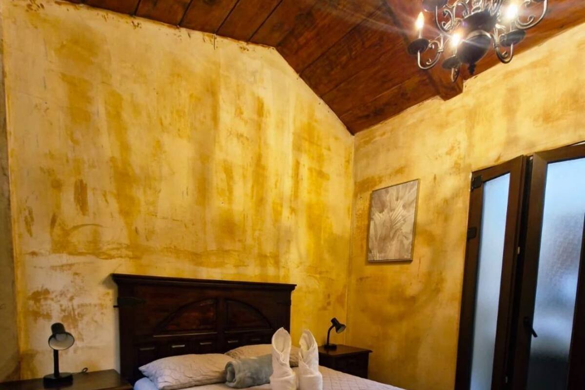 Une chambre avec un plafond en bois, un lit recouvert d’une courtepointe matelassée, une table de chevet, une lampe et un tableau accroché au mur.