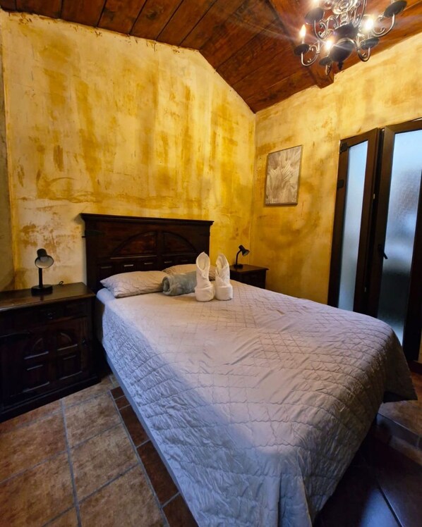 A bedroom with a wooden ceiling, a bed with a quilted bedspread, a nightstand, a lamp, and a painting on the wall.