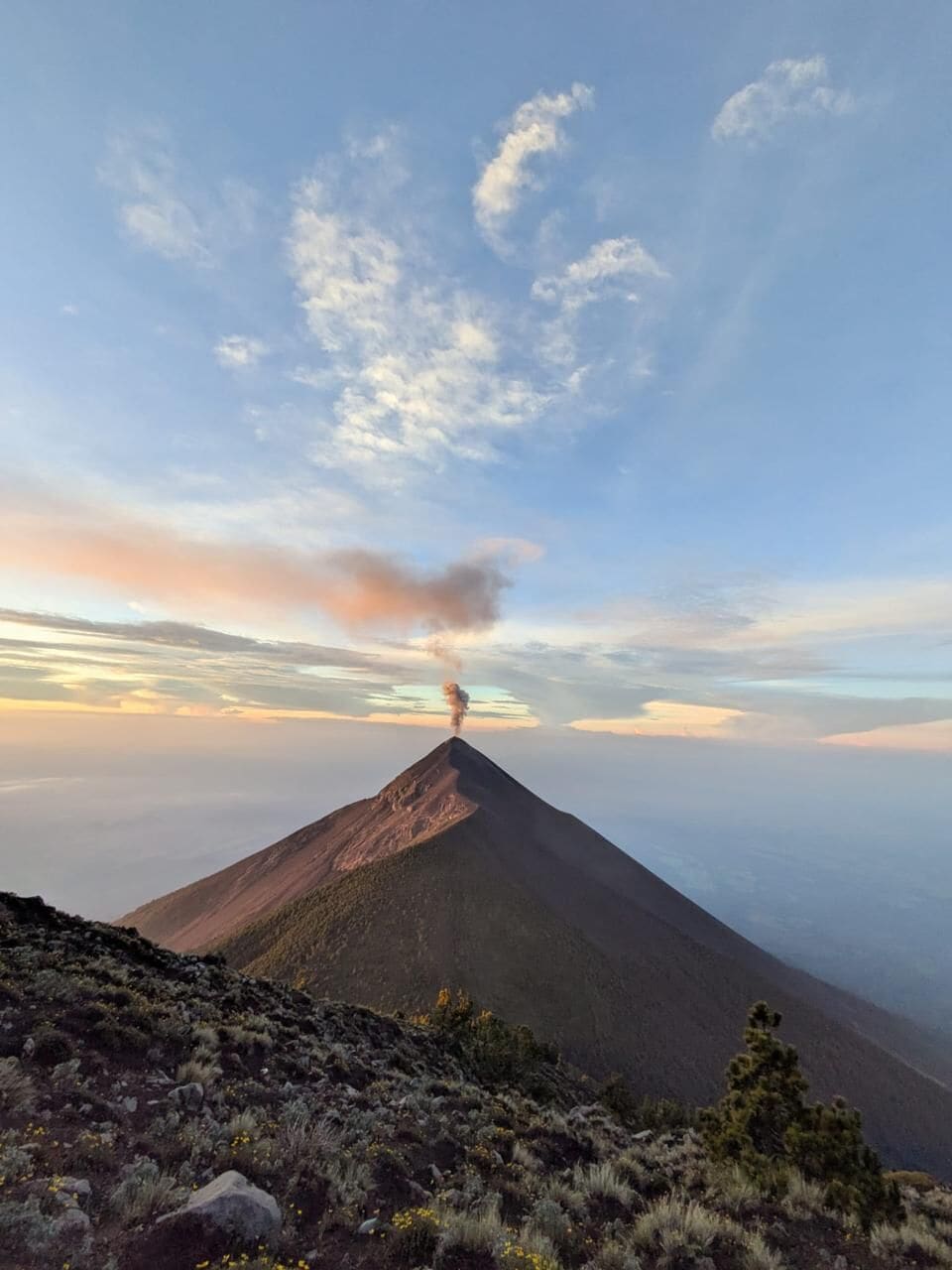 Una montaña con una columna de humo visible, rodeada de vegetación escasa y un cielo despejado.