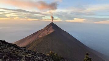 Una montaña con una columna de humo visible, rodeada de vegetación escasa y un cielo despejado.