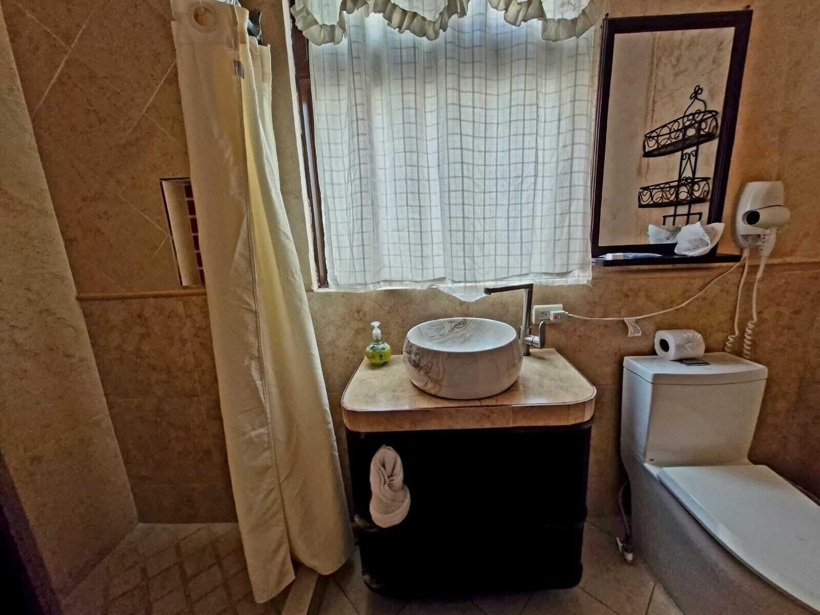 A bathroom with a toilet, a sink with a marble basin, a towel hanging on the wall, a shower curtain, and a window with a curtain.