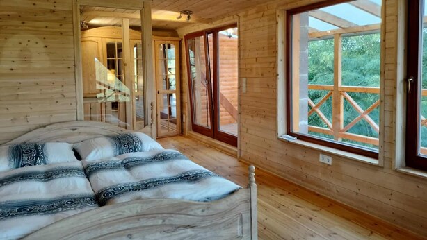 A wooden cabin bedroom with a bed, a window with a view of greenery, and a wooden staircase. - Wooden house in the middle of nature with a wonderful panorama, near to Budapest (Penc)