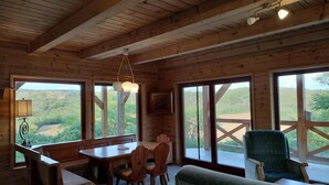 A wooden cabin interior with a dining table, chairs, a sofa, and large windows offering a view of the outdoors. - Wooden house in the middle of nature with a wonderful panorama, near to Budapest (Penc)