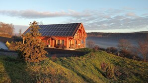 A wooden cabin with a sloped roof and solar panels, surrounded by a lush landscape and a body of water. - Wooden house in the middle of nature with a wonderful panorama, near to Budapest (Penc)