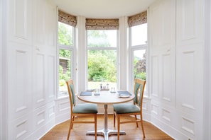 Une baie vitrée avec des murs à panneaux blancs, une table ronde en bois entourée de chaises, et une vue sur la verdure à l’extérieur.