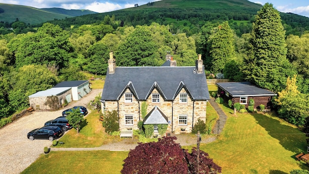 A stone building with a sloped roof, surrounded by lush greenery and hills. - Country House Near Loch Earn (Lochearnhead)