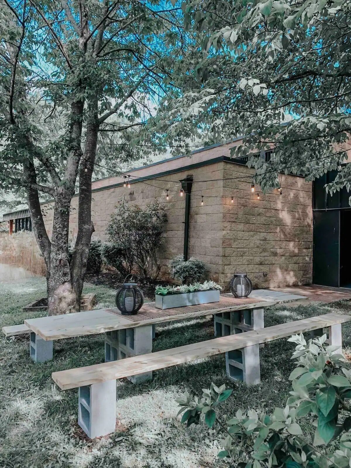 An outdoor patio area with a stone building, wooden benches, and string lights.