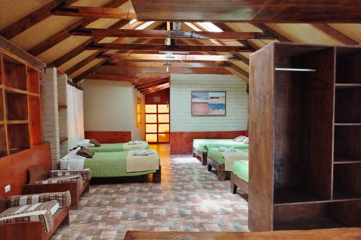 A room with wooden beams, a brick wall, and a tiled floor. There are two beds with green bedding and a wooden bench.