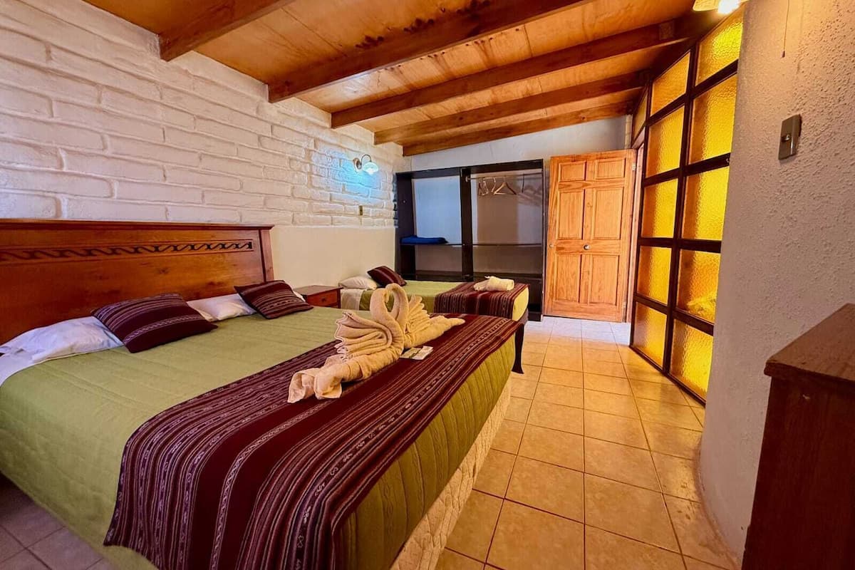 A hotel room with a wooden ceiling, a brick wall, a bed with a green bedspread, a wooden headboard, and a large window with frosted glass.