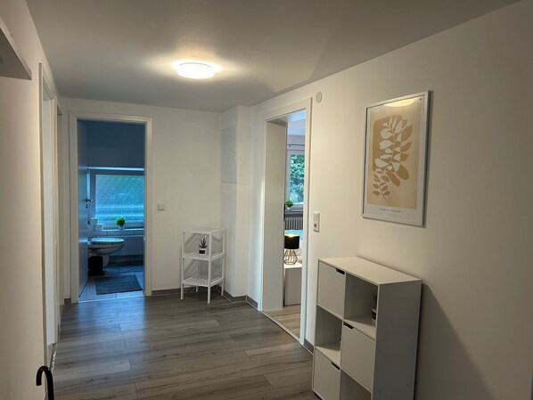 Un pasillo con piso de madera, paredes blancas y una lámpara en el techo. Se observa una obra de arte enmarcada en la pared y un estante blanco.