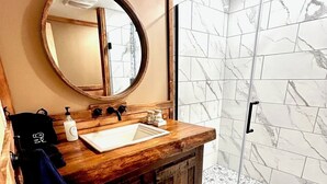 A bathroom with a wooden vanity, a round mirror, a sink, and a shower with marble tiles. - Stay in this historic Automotive-themed huge Apt. Located in charming Whitingham (Whitingham)