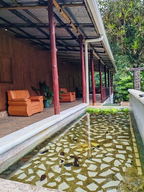 Een overdekt terras met twee oranje stoelen, een waterpartij met stenen tegels en een houten constructie met rode pilaren.