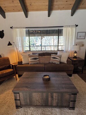 Ein gemütliches Wohnzimmer mit einem braunen Sofa, einem Holztisch und einem großen Fenster mit Vorhängen.