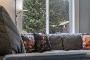 Un sofà amb coixinets múltiples, una finestra amb vista a arbres, i una làmpada a l'esquerra.
