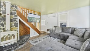 A living room with a stone wall, a wooden staircase, a grey sofa, and a television on the wall. - LOCATION~Walk 2 SeaLife&Parks~Perfect 4 Families (Seward)