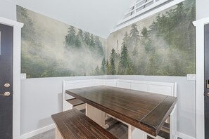 Huoneessa on tumma puinen pöytä ja penkki, valkoiset seinät ja seinämaalaus, jossa on kuvattuna metsä.