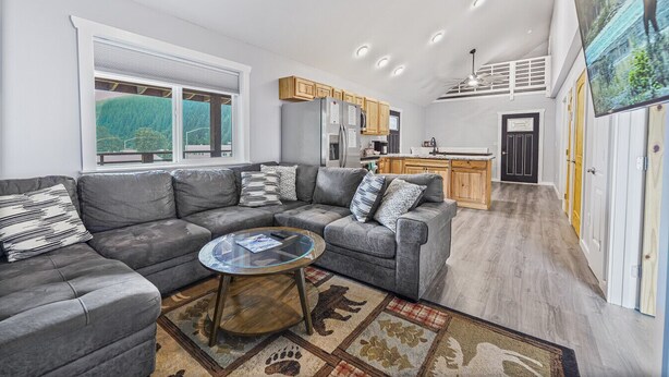 A modern living room with a grey sectional sofa, a glass coffee table, and a large window with a view of greenery. - The Wilders View | Downtown Seward · Deck Views · Sleeps 7 (Seward)