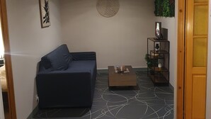 A living room with a dark blue sofa, a coffee table, and a patterned floor. There is a wall-mounted decoration and a shelf with items on the right side. - 100m2 apartment between lakes and mountains (Nantua)