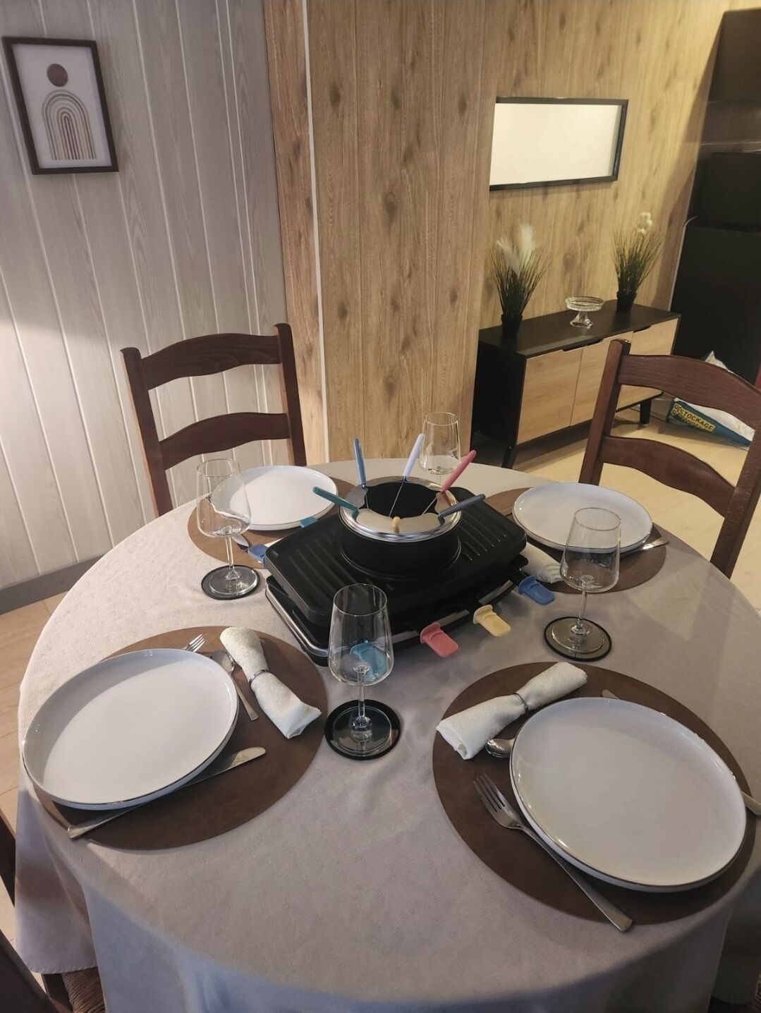 A round dining table with white plates, glasses, and cutlery, set for four. A black portable heater is in the center.