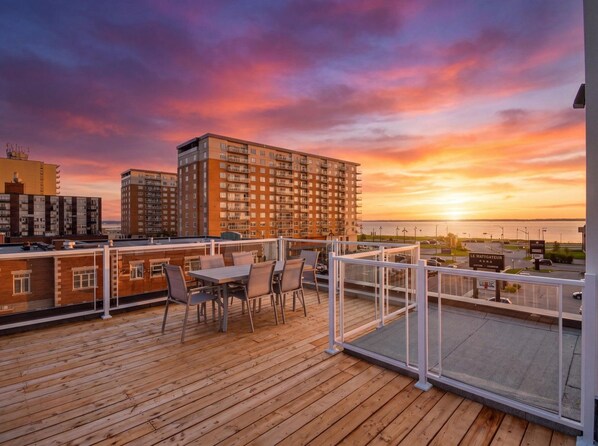 Una terraza de madera con mobiliario exterior y vistas a un atardecer sobre el horizonte de la ciudad.