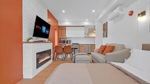 A modern living room with a fireplace, a flat-screen TV, a sofa, and a dining area with chairs. - Apartment 103 | NEW | Downtown (Rimouski)