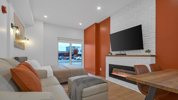A modern living room with a fireplace, a flat-screen TV, and a dining area with chairs. - Apartment 103 | NEW | Downtown (Rimouski)
