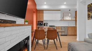 A modern kitchen with white cabinets, a wooden dining table, two chairs, a fireplace, and a kitchen island with a stove and microwave. - Apartment 103 | NEW | Downtown (Rimouski)