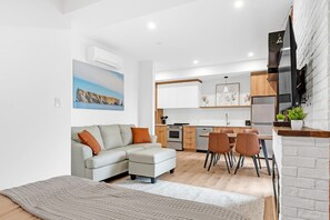 A modern living room with a sofa, a dining area with chairs, a kitchen with cabinets and appliances, and a large painting on the wall.
