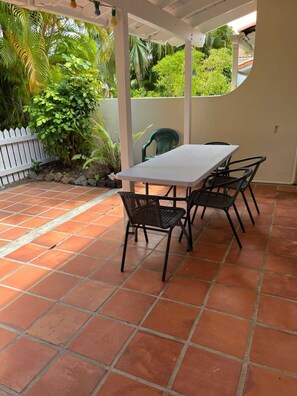 A covered patio with a white table and four black chairs, surrounded by terracotta tiles and lush greenery.