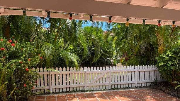 Un'area patio con recinto bianco a maglie, piastrelle in terracotta e rigogliosa vegetazione, tra cui palme e piante fiorite.