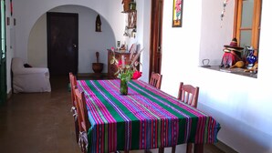 A dining area with a table covered in a colorful tablecloth, wooden chairs, a vase with flowers, and a hanging light fixture. - Finca historica los algarrobos (Salta)