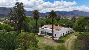 A white building with a tiled roof, surrounded by lush greenery and palm trees, set against a backdrop of mountains and a clear sky. - Finca historica los algarrobos (Salta)