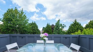 A glass-top table with white chairs and a vase of flowers on a rooftop terrace with a view of trees and a blue sky. - Surrey Family Home w/ Patio & Garden – 3BR 2BA (Surrey)