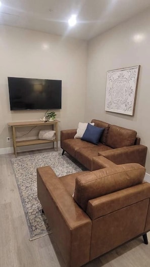 A living room with a brown sofa, a TV, a wooden TV stand, a patterned rug, and a framed artwork on the wall.