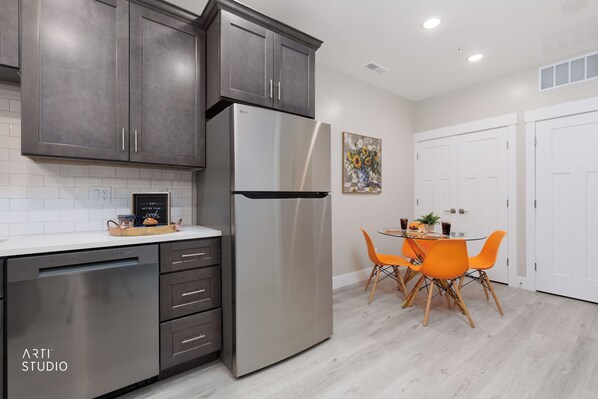 A modern kitchen with grey cabinets, a stainless steel refrigerator, a dishwasher, and a dining area with orange chairs.