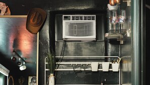 A compact kitchen with a refrigerator, a window with an air conditioner, and a shelf with glasses and a hat.