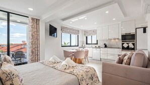 Una camera d'albergo moderna con un letto grande, una zona salotto con divano e poltrone, angolo cottura e un balcone vista città.