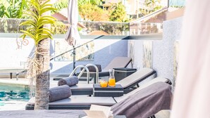 Uma área de descanso à beira da piscina, com espreguiçadeiras, uma palmeira e um livro.