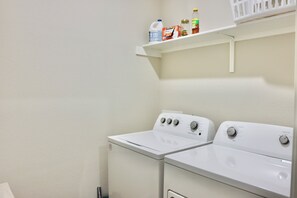 A laundry area with two white front-loading washing machines and a shelf above containing various bottles and containers. - Casa Encanto - Modern Stay, 2 Lounges, Gym & Fire Pit (Pflugerville)