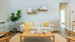 A living room with a sofa, coffee table, and a plant. There are framed pictures on the wall and a door leading to another room. - Casa Encanto - Modern Stay, 2 Lounges, Gym & Fire Pit (Pflugerville)