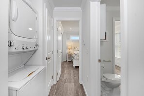 A laundry room with a front-loading washer and dryer, a hallway leading to a bedroom with a bed and nightstand, and a bathroom with a toilet and sink.