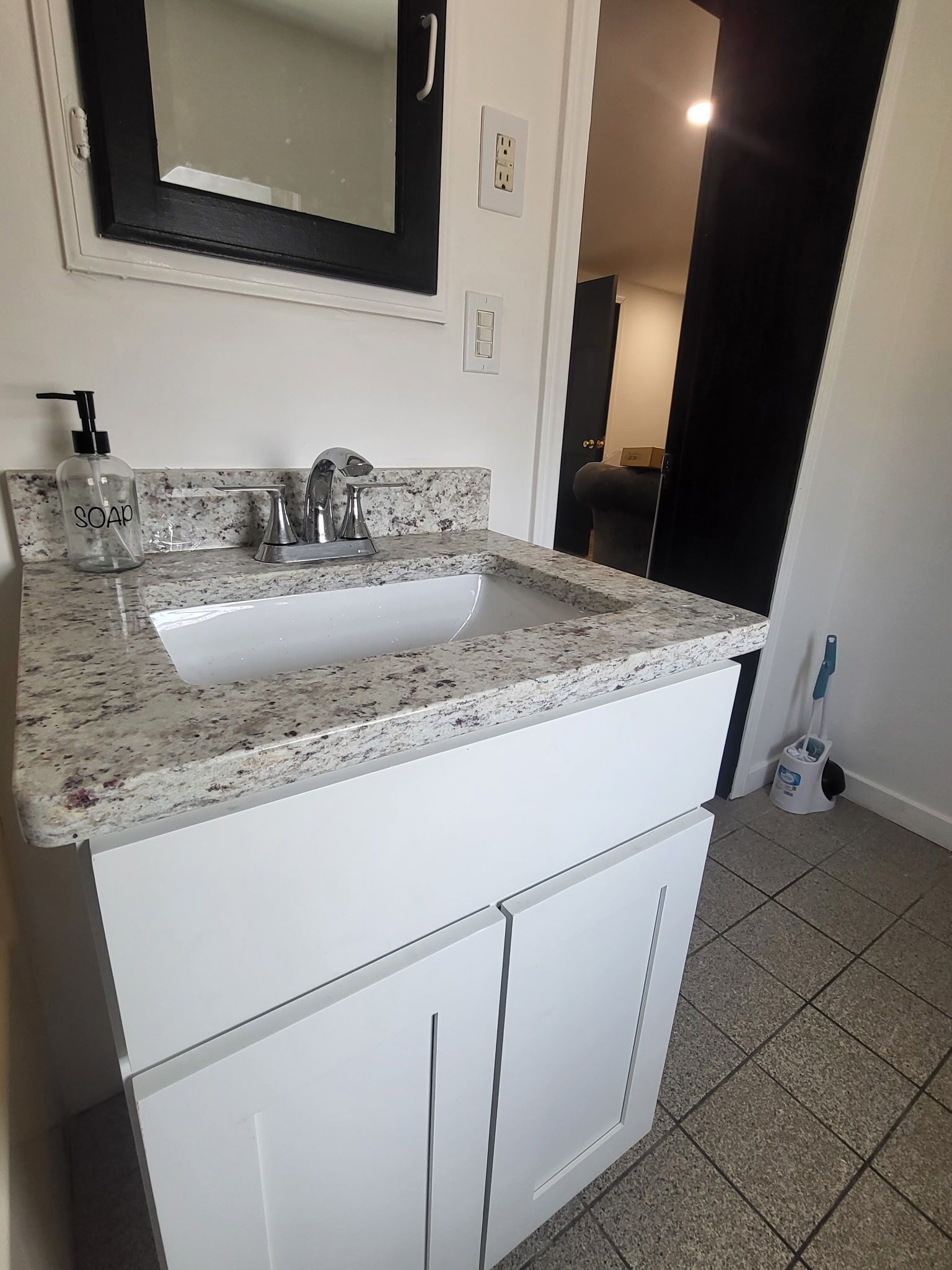 Une salle de bain avec un lavabo sur un plan de travail en granit, des meubles blancs, un miroir et une serpillière sur le sol.