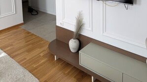 Ein modernes Wohnzimmer mit einem Flachbildfernseher an einer weißen Wand, einer hölzernen Bank, einer kleinen weißen Vase mit einer Feder und einem hellen Konsolentisch.