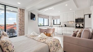 Une chambre d’hôtel moderne dotée d’un grand lit, d’un coin salon avec un canapé et des fauteuils, d’un coin cuisine et d’un balcon offrant une vue sur la ville.
