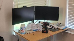 Un bureau avec deux écrans, des papiers, une boîte de Lyo et un outil électrique.